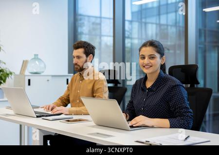 Multiracial business professionals are actively working on laptops at a shared desk in a contemporary open-plan office, highlighting teamwork and focused productivity in a modern workplace setting Stock Photo