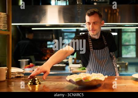 Caucasian young adult man wearing apron standing behind restaurant counter reaching to ring service bell, smiling while working in professional kitchen with food preparation visible Stock Photo