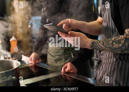 Young adult Caucasian man with tattooed arm using digital tablet in professional kitchen while another young adult man standing nearby, both engaged in culinary preparation near stove Stock Photo