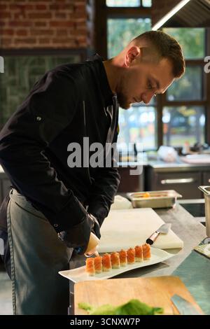 Caucasian young adult man preparing sushi rolls in professional kitchen, concentrating on arranging pieces on rectangular plate, wearing chef uniform, working at stainless steel counter Stock Photo