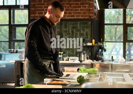 Caucasian young adult man preparing sushi rolls in professional kitchen, standing at counter with ingredients, wearing chef uniform and gloves, focused on culinary task Stock Photo