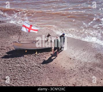 Sidmouth. An alsation dog sits in a small boat flying a St Georges flag on the beach at Sidmouth, Devon. Stock Photo