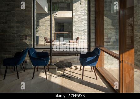 A sophisticated interior lounge space with plush blue chairs and a coffee table, separated by a large window from an outdoor patio with brown chairs. Stock Photo