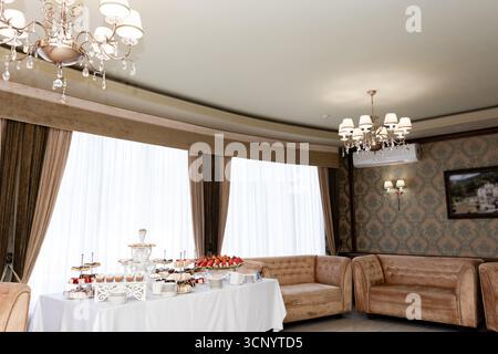 A lavish dessert spread is showcased on a white tablecloth, set against a backdrop of elegant furnishings and large windows, creating a sophisticated Stock Photo