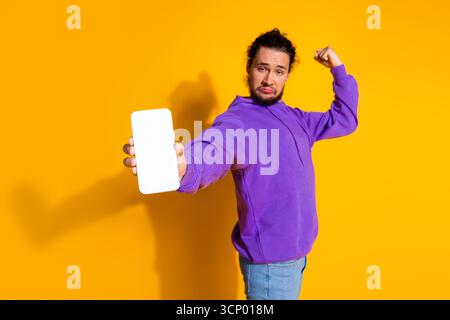 Young man in a purple hoodie displays a smartphone while making a confident bicep gesture, vibrant yellow background Stock Photo