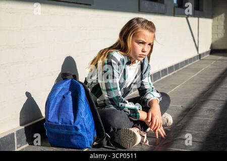 Female child sitting in school corridor by backpack with sunlight casting shadows, copy space Stock Photo