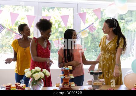 Diverse female friends gathering around table at home with tiered cupcakes and pink pennant banners Stock Photo