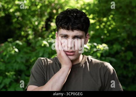 Worried young man holding face, his tooth hurts badly, he needs to go to the dentist. A young man appears thoughtful and concerned while outdoors Stock Photo