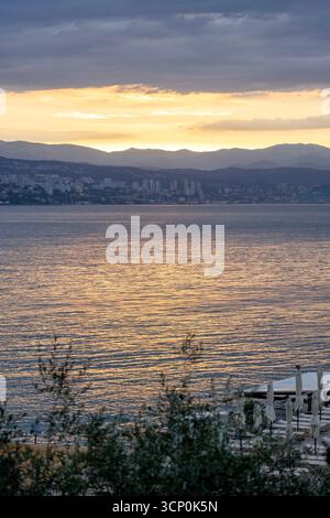 Golden sunset over calm sea with mountain silhouettes and distant city . Stock Photo