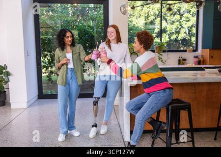 Diverse female friends toasting pink smoothies at marble island in kitchen under globe lights Stock Photo
