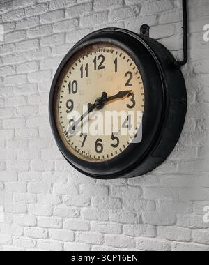 Old clock on white wall. Old analog railway clock with beige clock face and thick black bezel mounted on white brick wall. Stock Photo