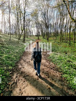 Walk in the woods - third person view. Female in jacket with backpack walking on a spring sunny day in a forest - view from the back. Stock Photo