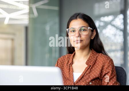 Confident young indian businesswoman wearing clear-framed glasses sitting at a desk in a modern office, portraying professionalism and focused determination Stock Photo