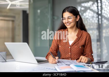 Young smiling indian woman wearing glasses and a dotted shirt, working intently at a desk with a laptop, writing notes and checking documents in a modern office environment Stock Photo