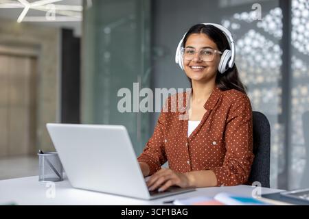 Young indian woman wearing white headphones and trendy glasses, working on a laptop at an office desk, smiling at the camera, representing remote work, education, and modern employee concepts Stock Photo