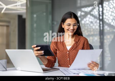 Young indian woman smiles while calculating finances, reviewing documents and using a laptop at a modern office desk, conveying confidence, focus and professional productivity Stock Photo