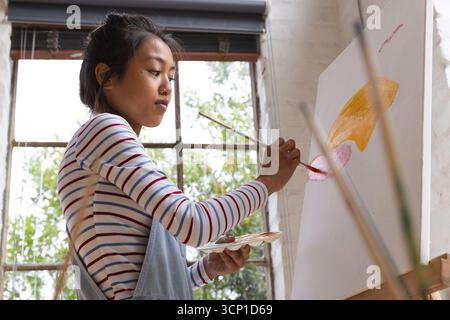 Asian woman in denim overalls applying orange and pink paint on canvas, holding palette in studio Stock Photo