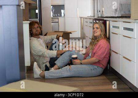 Couple holding disposable coffee cups while sitting on wood floor in kitchen with moving boxes Stock Photo