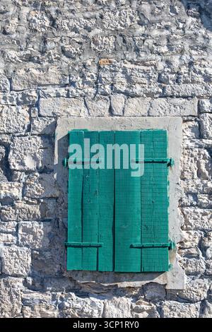 A weathered stone wall frames bold teal-green shutters, creating a rustic cottage-like facade. The textured surface and aged wood evoke history, durab Stock Photo