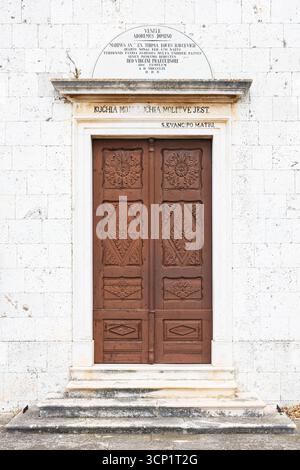 A weathered stone church with a bell tower and cross stands beside a sturdy stone wall. Green doors and rustic masonry evoke timeless rural architectu Stock Photo