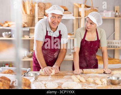 Older baker with younger assistant kneading and rolling dough in bakery Stock Photo