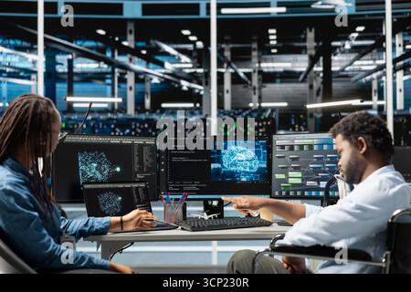 Data center engineer in wheelchair and coworker using AI visualization to oversee automated tasks. Worker with disability in inclusive server farm and colleague modifying systems based on analytics Stock Photo