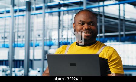 Happy engineer using laptop in industry 4.0 industrial plant to install machinery, conducting thorough testing. Cheerful robotics technician using notebook in smart factory calibrating IoT sensors Stock Photo