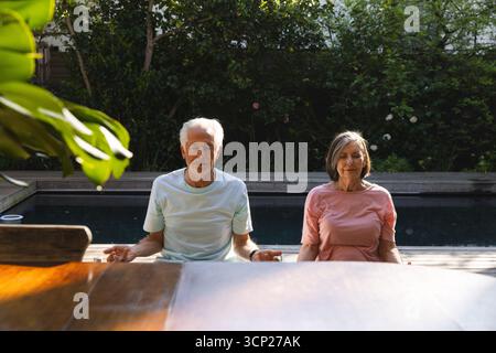 Senior couple meditating cross-legged on wooden pool deck by pool in backyard near table and shrubs Stock Photo