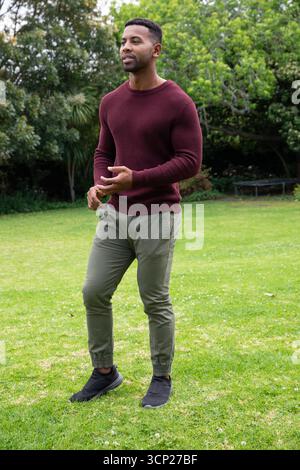 African american man standing on grassy lawn wearing burgundy knit sweater and jogger pants Stock Photo