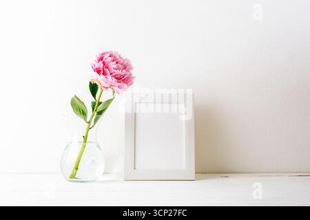 Minimal still life with pink peony in glass vase and blank picture frame on a table against white wall. Mockup. Stock Photo