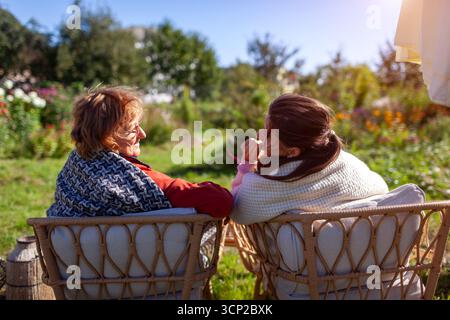 Senior woman chatting with her adult daughter relaxing in fall garden sitting on outdoor chairs covered with blankets. Back view. Family time Stock Photo
