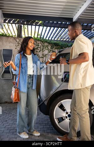 African American friends exchanging coffee cups while charging electric vehicle under metal carport Stock Photo