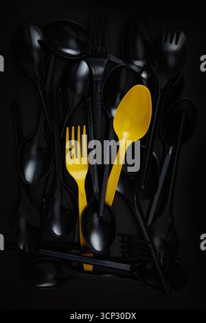 yellow plastic spoons and forks stand out among a pile of black plastic spoons and forks on a dark background Stock Photo
