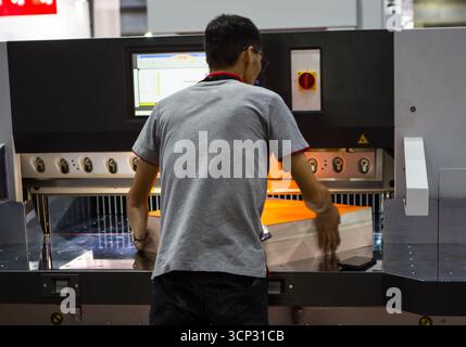 Back view of a technician adjusting large stacks of advertising paper on paper cutting machine. Stock Photo