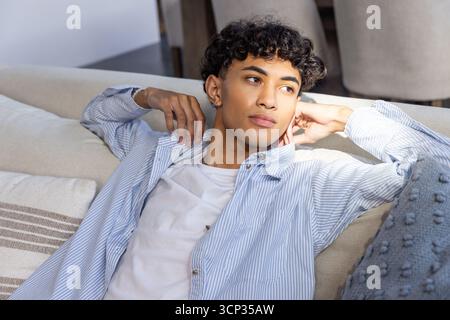 Asian man reclining on light-colored sofa in living room with pillows and dining chairs Stock Photo