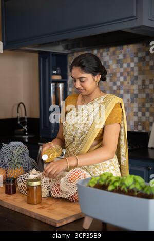 Indian woman in sari standing at kitchen counter while holding oil and reaching into mesh bag Stock Photo
