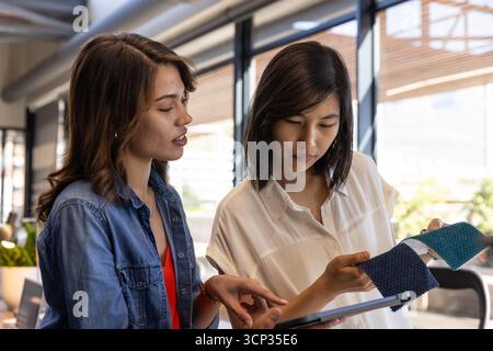 Diverse female colleagues collaborating on fabric swatch selection using tablet in office Stock Photo