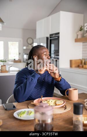 African american man sitting at table in kitchen eating scrambled eggs toast, pouring coffee Stock Photo