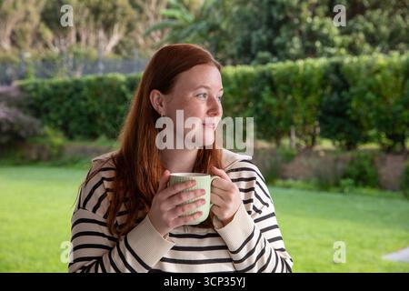 Adult redheaded woman standing on lawn near hedge holding green mug, looking pensively right Stock Photo