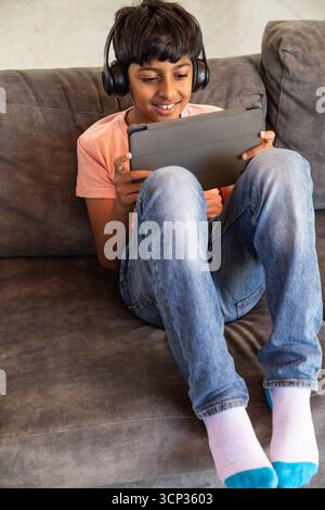Indian boy sitting on sofa at home using tablet with headphones near cushions Stock Photo