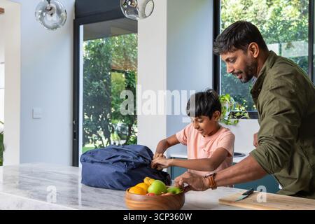 Indian father and son standing at marble island countertop unpacking blue backpack, copy space Stock Photo
