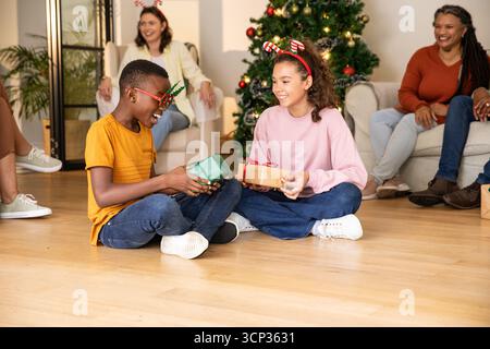 Diverse children wearing antler headbands exchanging gifts on floor at home, christmas tree Stock Photo