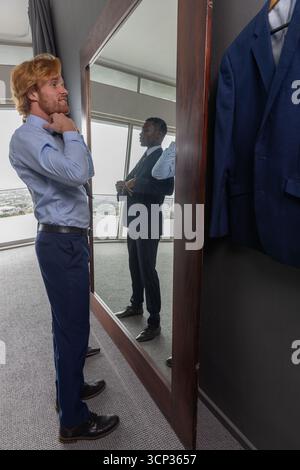 Diverse male colleagues adjusting formal attire in hotel suite with mirror, suit jacket Stock Photo