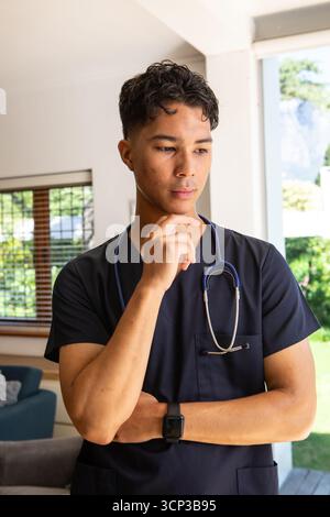 Asian male medical professional wearing scrubs, thinking in living room holding stethoscope Stock Photo