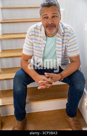Senior Hispanic man leaning forward with clasped hands on wooden staircase at home Stock Photo