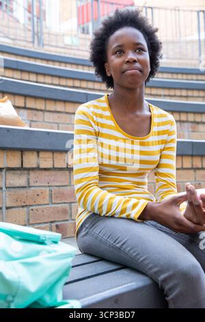 Adolescent african american female student sitting on bench at campus quad next to backpack Stock Photo