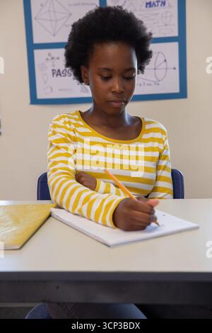 African american girl writing in notebook with pencil in classroom wearing striped shirt Stock Photo