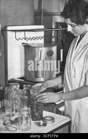 A lady with various glass jars on an enamel worktop. Behind her is a tall jam making pot with a thermometer, on a Vulcan cooker. From a printed photograph which originally appeared in the publication, The Vulcan Cook Book, which was published in 1936 by the Vulcan Stove Company of Exeter, England. Stock Photo