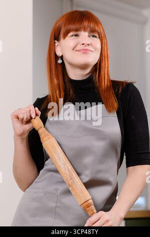 Portrait of happy smiling female cook, housewife, baker in kitchen apron, holding rolling pin, cooking in kitchen. Culinary concept Stock Photo