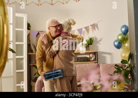 Senior Man Hugging Senior Woman While Holding Gift Box Stock Photo